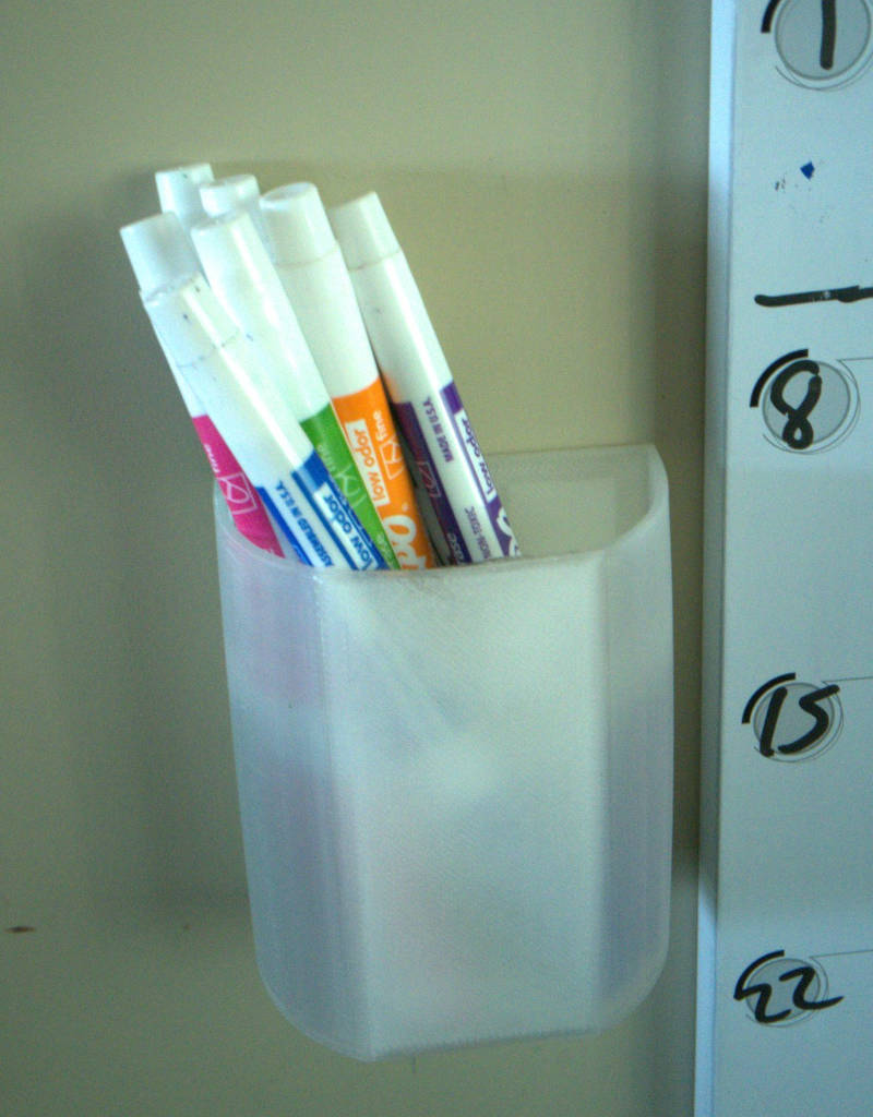 A D-shaped, open-top container affixed to a wall holding dry-erase pens next to the edge of a dry-erase calendar.